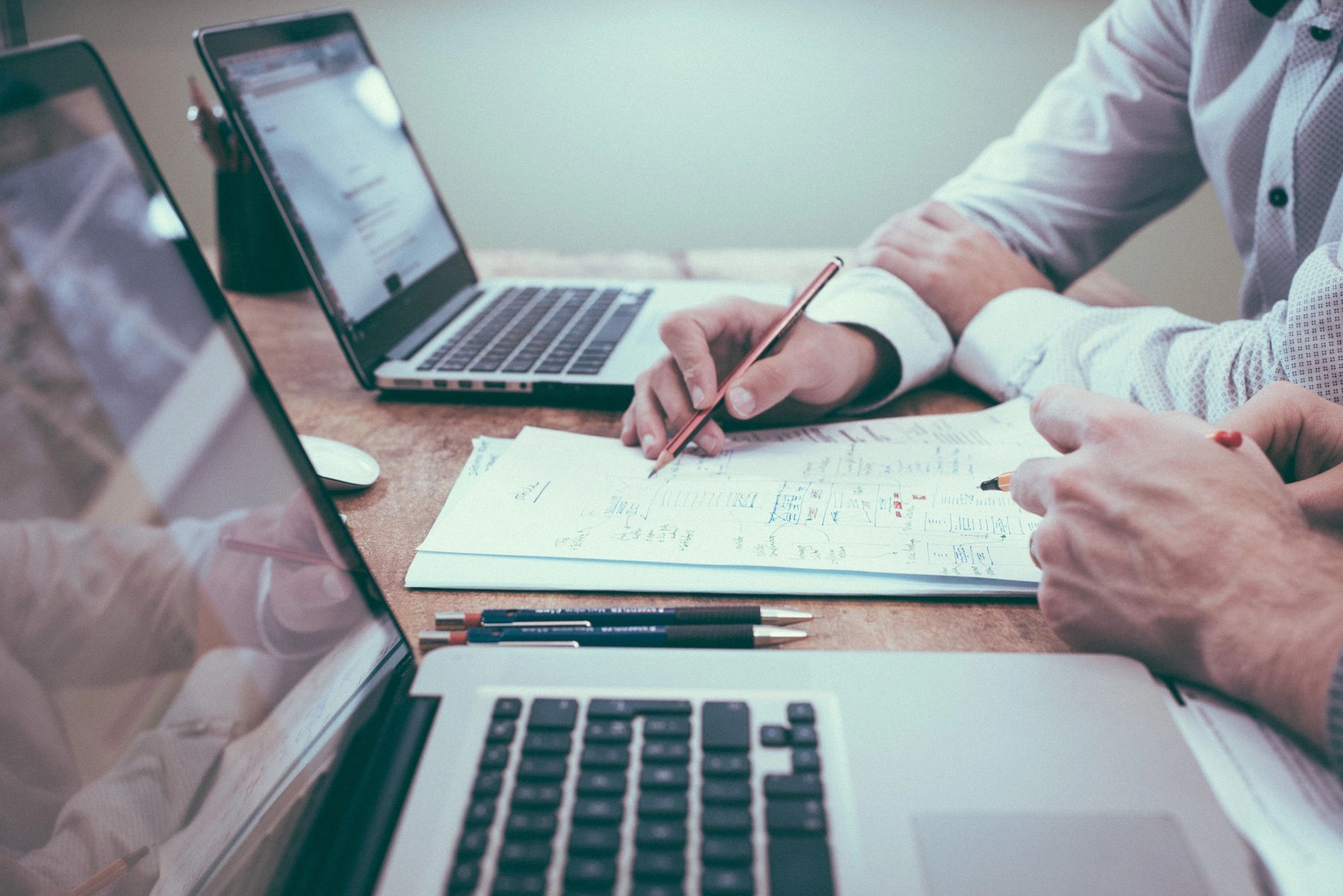 Two people collaborating over laptops and documents on a wooden table.